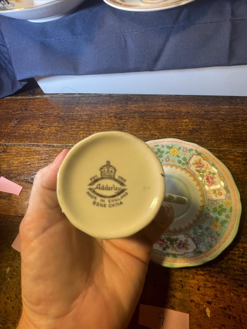 Może przedstawiać: Zestaw do herbaty z porcelany kostnej w stylu vintage z kwiatowym spodkiem. Na spodzie filiżanki widnieje napis "Adderley Made in England Bone China" i logo korony. Spodek ma zielony i r&oacute;żowy wz&oacute;r kwiatowy.