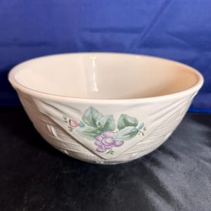 May include: A white ceramic bowl with a woven texture and a grape design. The bowl has a wide, shallow shape and a smooth interior. The grape design features purple grapes and green leaves.