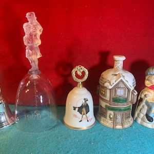 May include: Five decorative bells with various designs. A clear glass bell with a figurine of a man carrying a child, a silver bell with a figurine of a man holding a staff, a white bell with a wreath design, a white bell with a man in a red coat, and a brown ceramic bell with a house design.