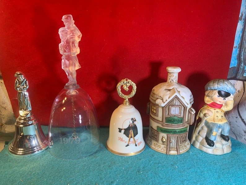 May include: Five decorative bells with various designs. A clear glass bell with a figurine of a man carrying a child, a silver bell with a figurine of a man holding a staff, a white bell with a wreath design, a white bell with a man in a red coat, and a brown ceramic bell with a house design.