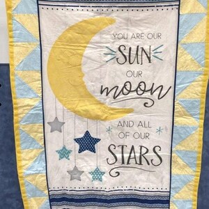 May include: A rectangular quilt featuring a central white panel with a yellow crescent moon and hanging stars. The text reads: YOU ARE OUR SUN OUR moon AND ALL OF OUR STARS. The border consists of yellow and light blue triangles with a yellow binding.