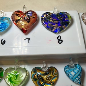 May include: Heart-shaped glass pendants in various colors. One pendant has a red and gold design, another has blue and gold, and others feature green, gold, and blue swirls. Each pendant has a clear glass loop for hanging.