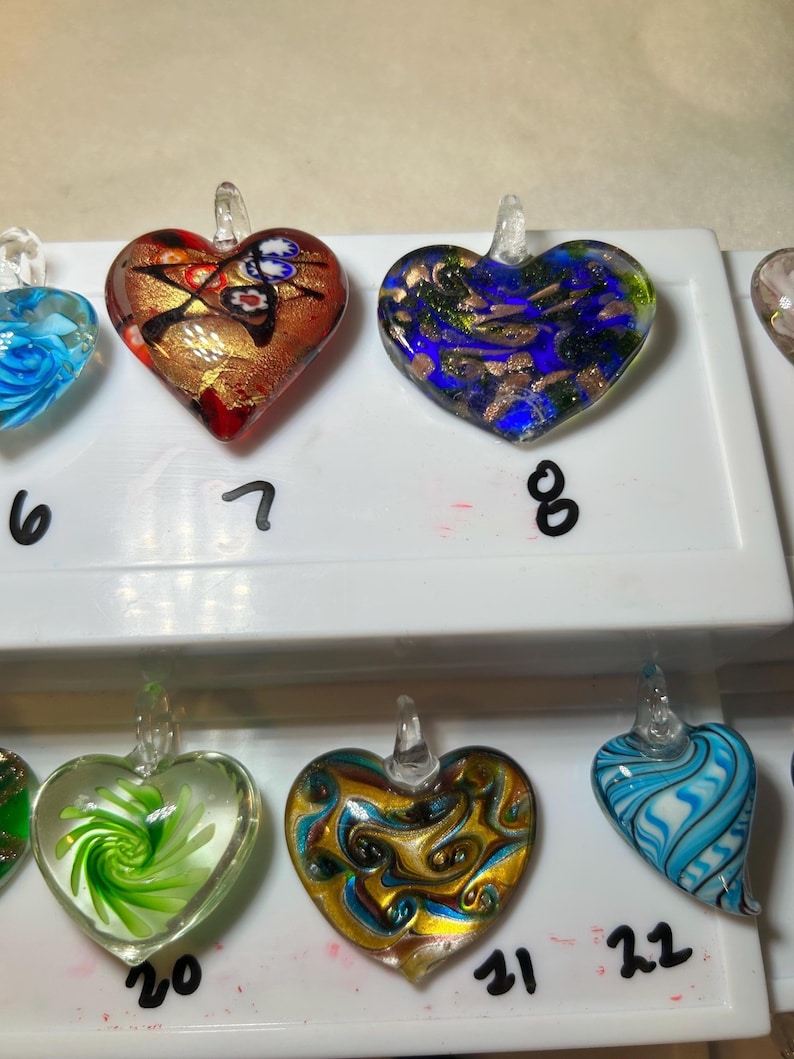 May include: Heart-shaped glass pendants in various colors. One pendant has a red and gold design, another has blue and gold, and others feature green, gold, and blue swirls. Each pendant has a clear glass loop for hanging.