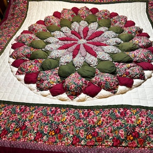 May include: A biscuit quilt featuring a central circular medallion of stuffed fabric puffs in floral, solid olive green, and solid burgundy patterns. The quilt has a white quilted border and a wide outer frame of floral fabric with burgundy trim.