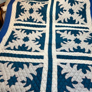May include: A handmade quilt with a striking blue and white design. The quilt features a repeating pattern of stylized floral and star motifs in blue against a white background. The quilt is bordered with a blue edge.