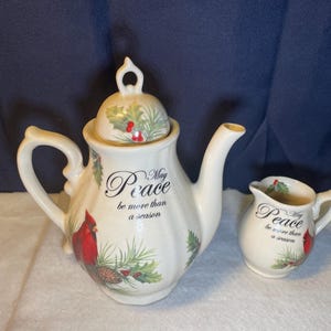 Puede incluir: Un juego de tetera y crema de cerámica color crema. La tetera tiene tapa y asa curva. Ambas piezas están decoradas con un cardenal rojo, agujas de pino y las palabras "May Peace be more than a season."