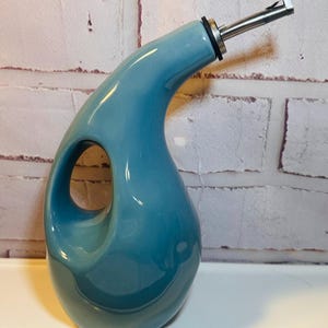 Puede incluir: Una aceitera de cerámica en color azul cerceta brillante con un asa ergonómica curva integrada en el cuerpo. El recipiente presenta un cuello largo y cónico rematado con un vertedor de acero inoxidable que incluye una junta de goma negra.