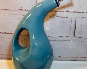 Puede incluir: Una aceitera de cerámica en color azul cerceta brillante con un asa ergonómica curva integrada en el cuerpo. El recipiente presenta un cuello largo y cónico rematado con un vertedor de acero inoxidable que incluye una junta de goma negra.