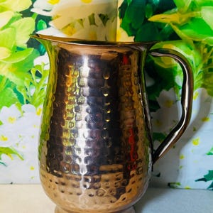 May include: A hammered copper pitcher with a curved handle and a wide spout. The pitcher has a textured surface with a series of small, evenly spaced indentations. The pitcher is designed to hold liquids and is suitable for serving beverages.
