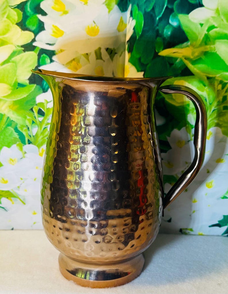 May include: A hammered copper pitcher with a curved handle and a wide spout. The pitcher has a textured surface with a series of small, evenly spaced indentations. The pitcher is designed to hold liquids and is suitable for serving beverages.