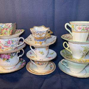 Op de afbeelding: Verschillende stapels vintage theekopjes en schotels. De kopjes hebben bloempatronen in verschillende kleuren, waaronder roze, geel en blauw. Sommige kopjes hebben gouden randen. De schotels hebben ook bloemmotieven en gouden randen.