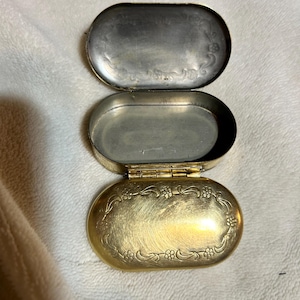 May include: An open, antique, oval-shaped pillbox with a gold-colored exterior and a silver-colored interior. The lid and base are decorated with floral patterns. The pillbox is hinged and appears to be made of metal.