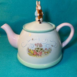 May include: A white ceramic teapot with a light pink handle and a green rim. The teapot features a floral design with a small rabbit figurine on the lid holding a sign that says "Peter Rabbit".