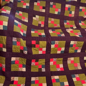 Könnte beinhalten: Handgefertigte Steppdecke mit Patchwork-Design. Die Steppdecke hat einen dunkelvioletten Hintergrund mit einem Gittermuster aus olivgrünen Quadraten. Jedes Quadrat enthält kleinere Quadrate aus rotem, blauem und gemustertem Stoff. Die Steppdecke ist ein gemütlicher, handgefertigter Artikel.