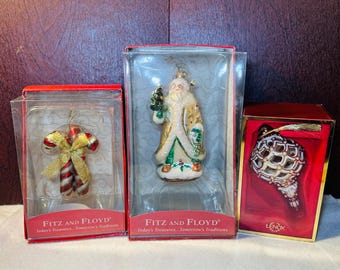 May include: Three Christmas ornaments in boxes. One is a candy cane with a gold bow, another is a Santa Claus figure, and the third is a white and gold teardrop-shaped ornament. The boxes have the text "Fitz and Floyd" and "Lenox".