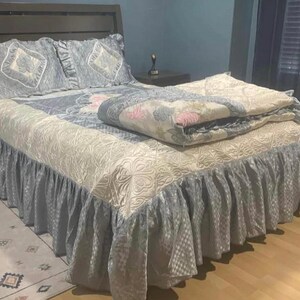 May include: A bed with a dark brown headboard, adorned with a matching comforter set. The set includes two decorative pillows, a patterned quilt, and a bed skirt in shades of cream and blue. The bed is placed on a light brown wooden floor.