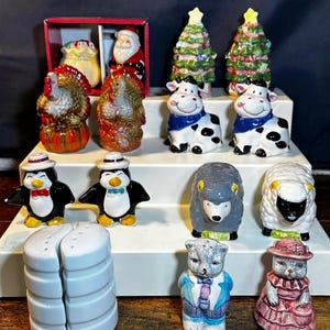 Puede incluir: Una colección de saleros y pimenteros de cerámica con varios diseños que incluyen pavos, vacas, pingüinos, ovejas, gatos y árboles de Navidad, junto con un set de Santa Claus en caja y un set cilíndrico blanco sobre un expositor escalonado.