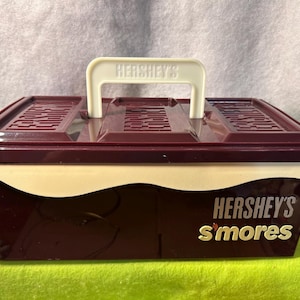 May include: A rectangular Hershey's S'mores container with a burgundy body and a cream-colored band. The lid has a burgundy top with a cream-colored handle. The words "HERSHEY'S S'mores" are printed on the front in silver and gold.