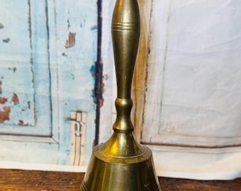 Pode incluir: Um sino de latão vintage com uma longa alça e uma base alargada. O sino tem um acabamento polido e é provavelmente usado para sinalização ou decoração. A alça apresenta um design decorativo e o sino fica em uma superfície de madeira.