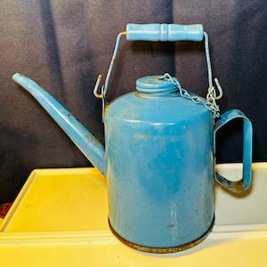 May include: A vintage, light blue metal watering can with a long spout and a handle. The can has a matching blue handle and a chain attached to the lid. The can shows signs of wear and rust, suggesting age and use. The can is sitting on a yellow surface.
