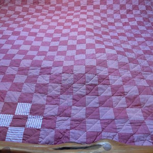 May include: A handmade quilt with a checkerboard pattern in shades of burgundy and white. The quilt features alternating squares of solid burgundy and burgundy and white striped fabric. The quilt is on a wooden bed frame.