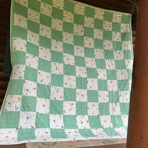 May include: A handmade quilt with a checkerboard pattern of white squares with floral designs and light green squares. The quilt is hanging on a wooden structure, showcasing its design and craftsmanship.