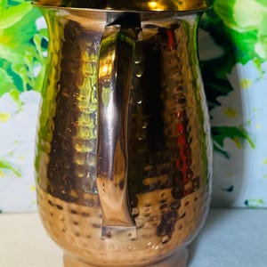 May include: A copper-colored pitcher with a hammered texture. The pitcher has a curved handle and a spout for pouring. The base is round and the body of the pitcher has a bulbous shape. The pitcher is made of metal and has a shiny finish.