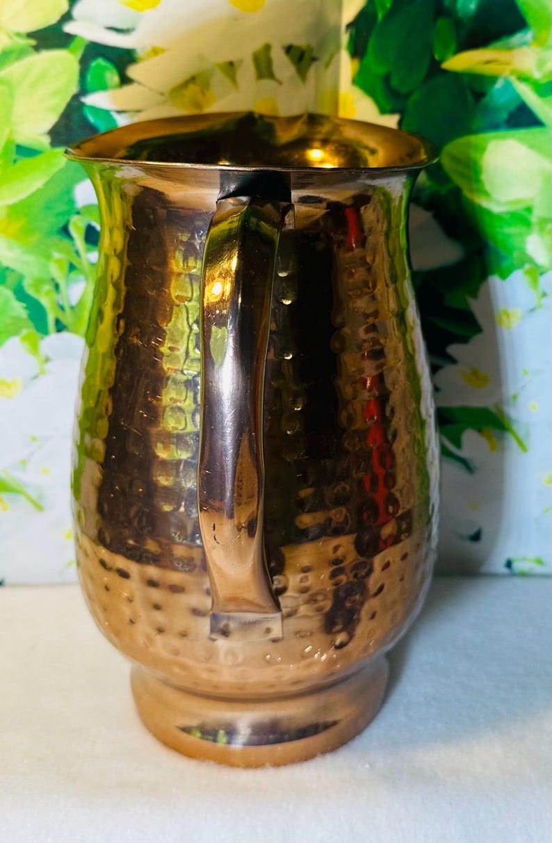 May include: A copper-colored pitcher with a hammered texture. The pitcher has a curved handle and a spout for pouring. The base is round and the body of the pitcher has a bulbous shape. The pitcher is made of metal and has a shiny finish.