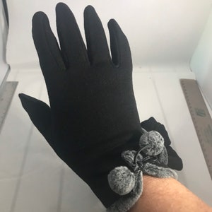 May include: Black gloves with a decorative cuff featuring gray faux fur trim and two gray pom-poms. The gloves are made of a smooth, stretchy material. The cuff is secured with a black metal ring.