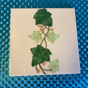 Puede incluir: Un azulejo de cerámica blanco con un diseño de hiedra verde y amarilla. La hiedra está pintada en un estilo realista con hojas verdes y bayas amarillas.