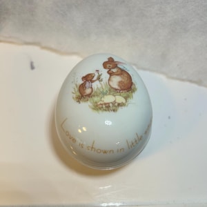 Peut inclure: Boîte à bijoux en céramique blanche en forme d'œuf avec une illustration peinte d'un lapin et d'une souris bruns. Le lapin et la souris sont dans un champ herbeux. Le texte "Love is shown in little ways" est écrit autour de la base de l'œuf.