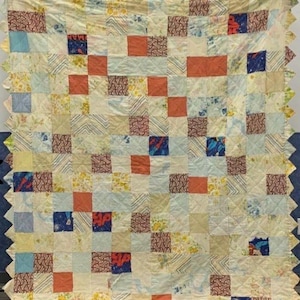 Puede incluir: Un edredón de retazos con un fondo color crema y un patrón de cuadrícula de cuadrados. Los cuadrados presentan varios patrones y colores, incluyendo estampados naranjas, azules y florales. El edredón tiene un borde festoneado.