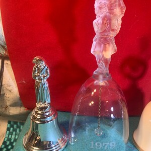 May include: Two vintage glass bells featuring frosted figures of people. The larger bell is clear glass with a frosted figure of a man and woman holding hands. The smaller bell is silver with a frosted figure of a man holding a staff. The larger bell has the year 1979 etched on the bottom.