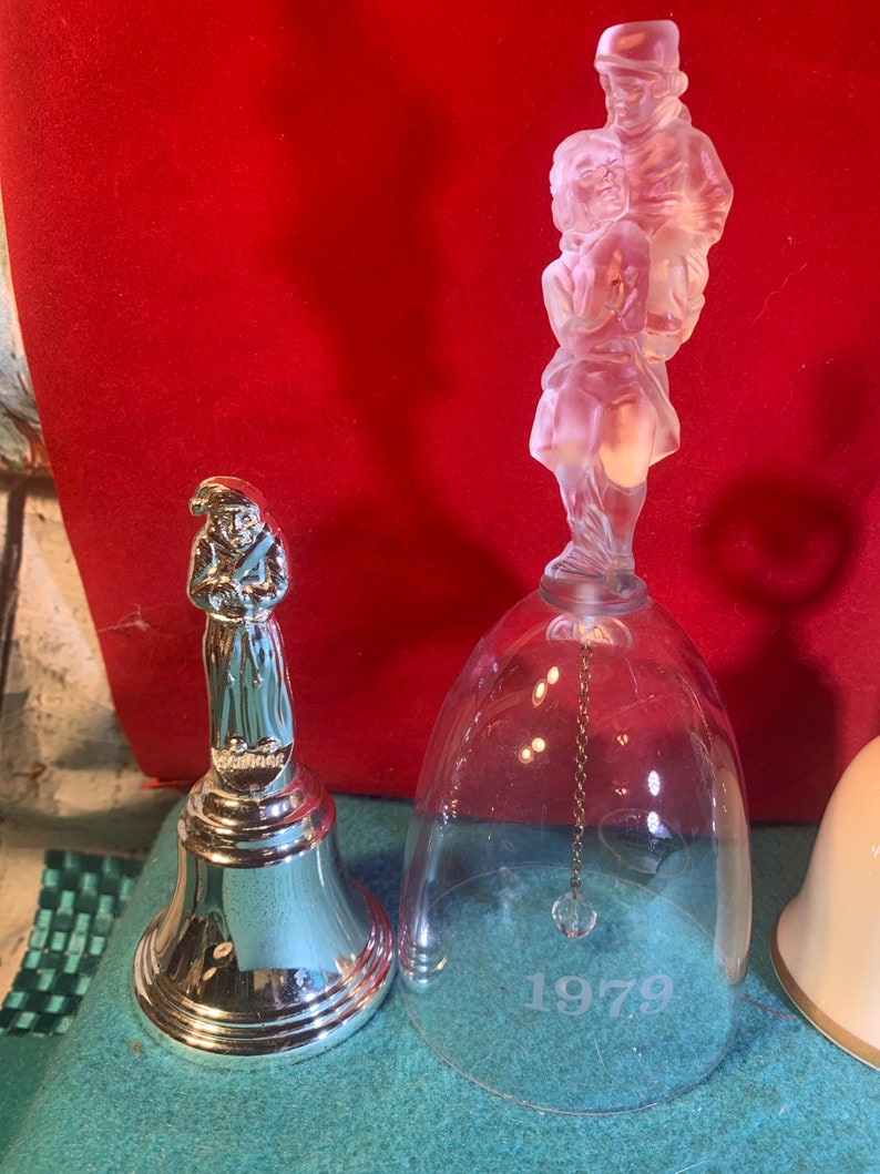 May include: Two vintage glass bells featuring frosted figures of people. The larger bell is clear glass with a frosted figure of a man and woman holding hands. The smaller bell is silver with a frosted figure of a man holding a staff. The larger bell has the year 1979 etched on the bottom.