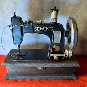 May include: A black vintage sewing machine with the word "SEWING" in gold lettering. The machine sits on a wooden base with a small latch. The sewing machine has a silver wheel and gold accents.
