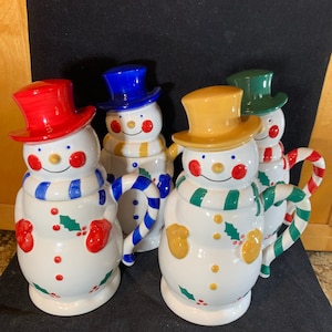 May include: Four ceramic snowman mugs with candy cane handles. Each mug has a different colored hat: red, blue, yellow, and green. The snowmen have white faces with red cheeks and black eyes.
