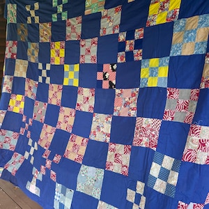 Può includere: Una trapunta patchwork caratterizzata da un motivo a griglia ripetuto di quadrati blu reale a tinta unita che si alternano a blocchi più piccoli a nove riquadri. I blocchi sono composti da vari tessuti floreali, a righe e geometrici.