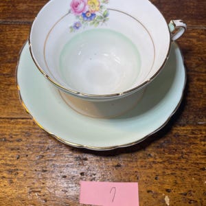 Może przedstawiać: Zestaw filiżanki i spodka do herbaty w stylu vintage. Filiżanka jest biała z kwiatowym wzorem w kolorze r&oacute;żowym, niebieskim i ż&oacute;łtym. Wnętrze filiżanki ma jasnozielony odcień. Spodek i rant filiżanki są jasnozielone ze złotym wykończeniem.