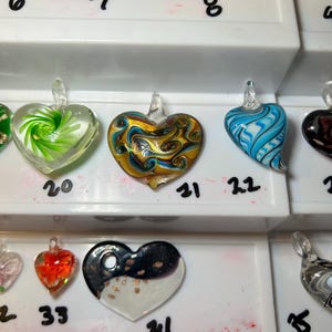 May include: A collection of heart-shaped glass pendants in various colors and designs. The pendants include swirls, patterns, and color combinations such as gold and blue, red and orange, and black and white. Each pendant has a small loop for hanging.