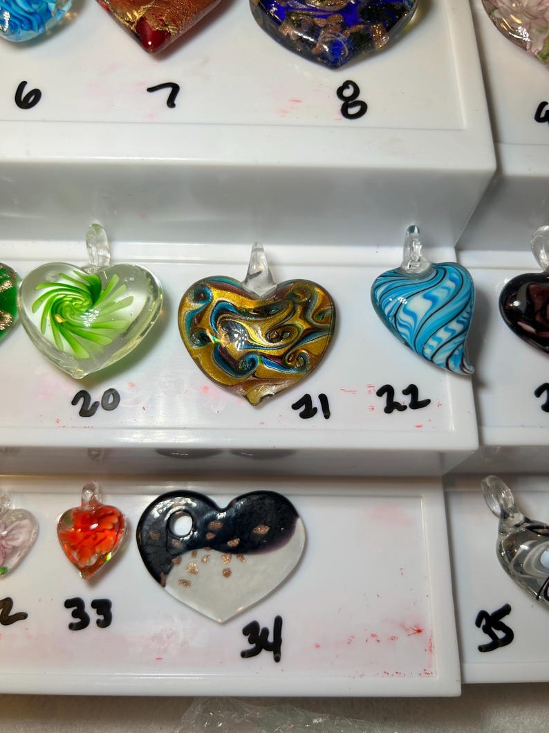 May include: A collection of heart-shaped glass pendants in various colors and designs. The pendants include swirls, patterns, and color combinations such as gold and blue, red and orange, and black and white. Each pendant has a small loop for hanging.