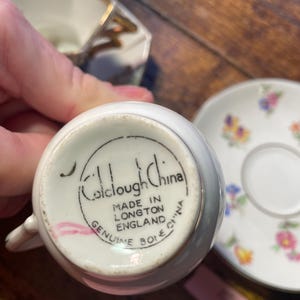Może przedstawiać: Zbliżenie białej filiżanki do herbaty z uchwytem, spodkiem i śmietanką. Na spodzie filiżanki widnieje napis "Colclough China" i "Made in Longton England". Spodek ma kwiatowy wz&oacute;r.