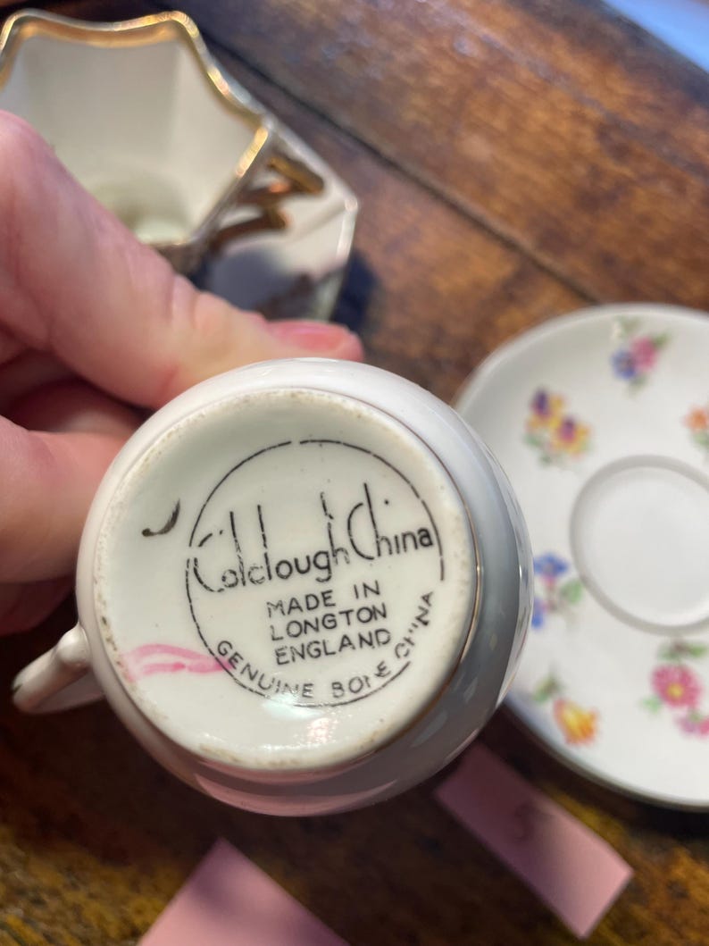 Może przedstawiać: Zbliżenie białej filiżanki do herbaty z uchwytem, spodkiem i śmietanką. Na spodzie filiżanki widnieje napis "Colclough China" i "Made in Longton England". Spodek ma kwiatowy wz&oacute;r.
