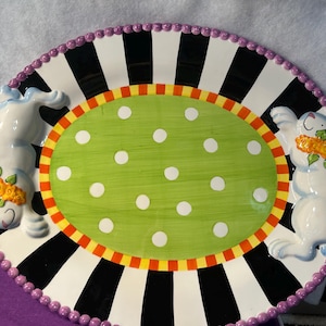 May include: Oval ceramic serving platter with a whimsical Easter theme. The platter features a green center with white polka dots, surrounded by a black and white striped border. Two white bunny figures with floral accents are on the sides.