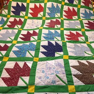 May include: A handmade quilt featuring a repeating pattern of tulip shapes in red, blue, and patterned fabrics. The tulips are set against a white background, framed by green and yellow borders. The quilt is likely made of cotton.