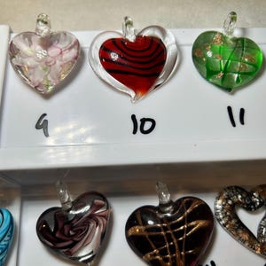 May include: A collection of heart-shaped glass pendants in various colors and designs. The pendants include floral patterns, red and black swirls, green with gold accents, and dark with gold lines. Each pendant has a small loop at the top.