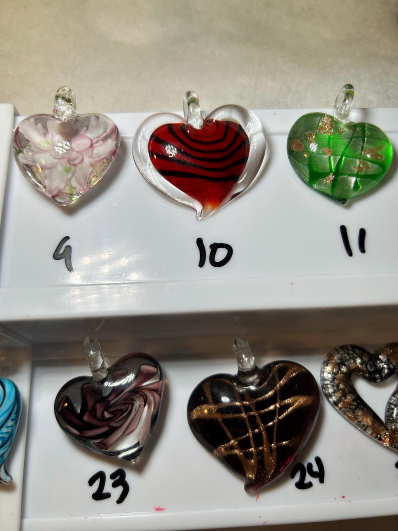 May include: A collection of heart-shaped glass pendants in various colors and designs. The pendants include floral patterns, red and black swirls, green with gold accents, and dark with gold lines. Each pendant has a small loop at the top.