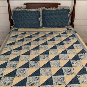 以下が含まれることがあります： 濃い木製のヘッドボードと、青とクリーム色の三角形の繰り返し模様が特徴のキルトが置かれたベッド。フリルエッジの付いたお揃いの青い模様の枕が2つ、ベッドの上に置かれています。