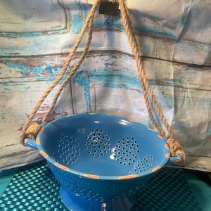 May include: A blue ceramic colander with a distressed finish, suspended by a natural rope and a binder clip. The colander has a pedestal base and numerous drainage holes. The rope is attached to the colander's handles.