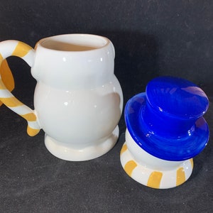 May include: A white ceramic snowman mug with a yellow and white candy cane handle. The mug is separated from the snowman's head, which is white with a blue hat and yellow and white stripes around the bottom.