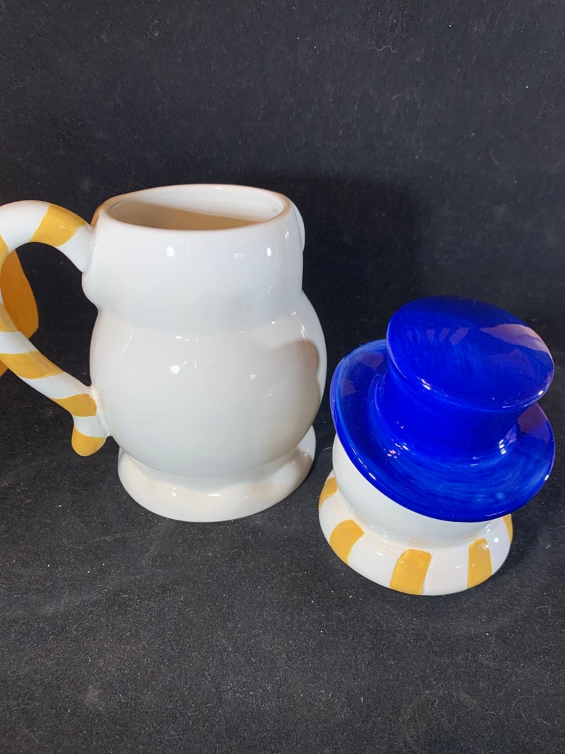 May include: A white ceramic snowman mug with a yellow and white candy cane handle. The mug is separated from the snowman's head, which is white with a blue hat and yellow and white stripes around the bottom.